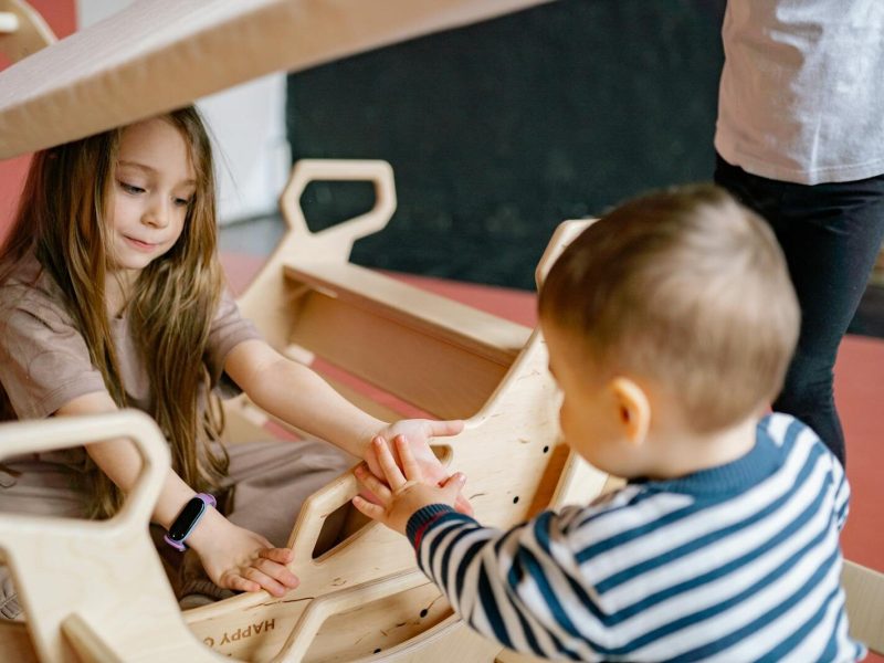 Children enjoying a playful moment with a wooden rocker indoors, emphasizing fun and interaction.